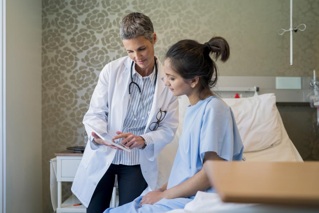 Patient in hospital gown on bed with doctor showing something on clipboard