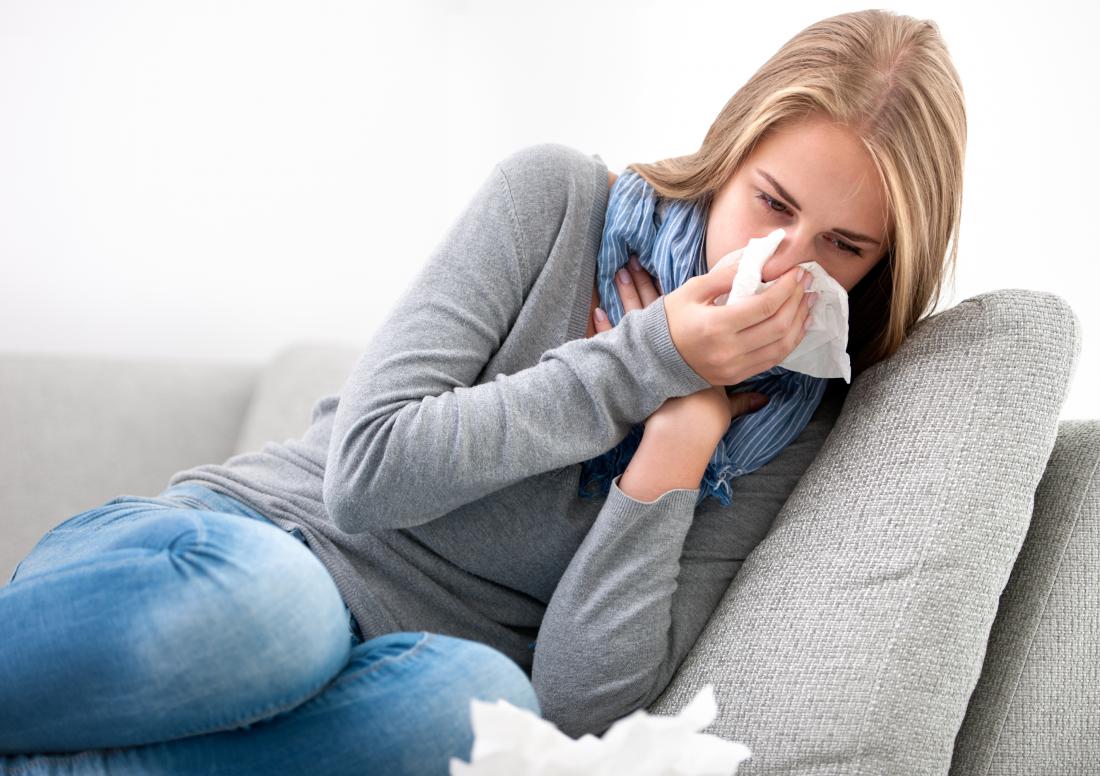 Woman unwell with infection blowing her nose sitting on sofa.