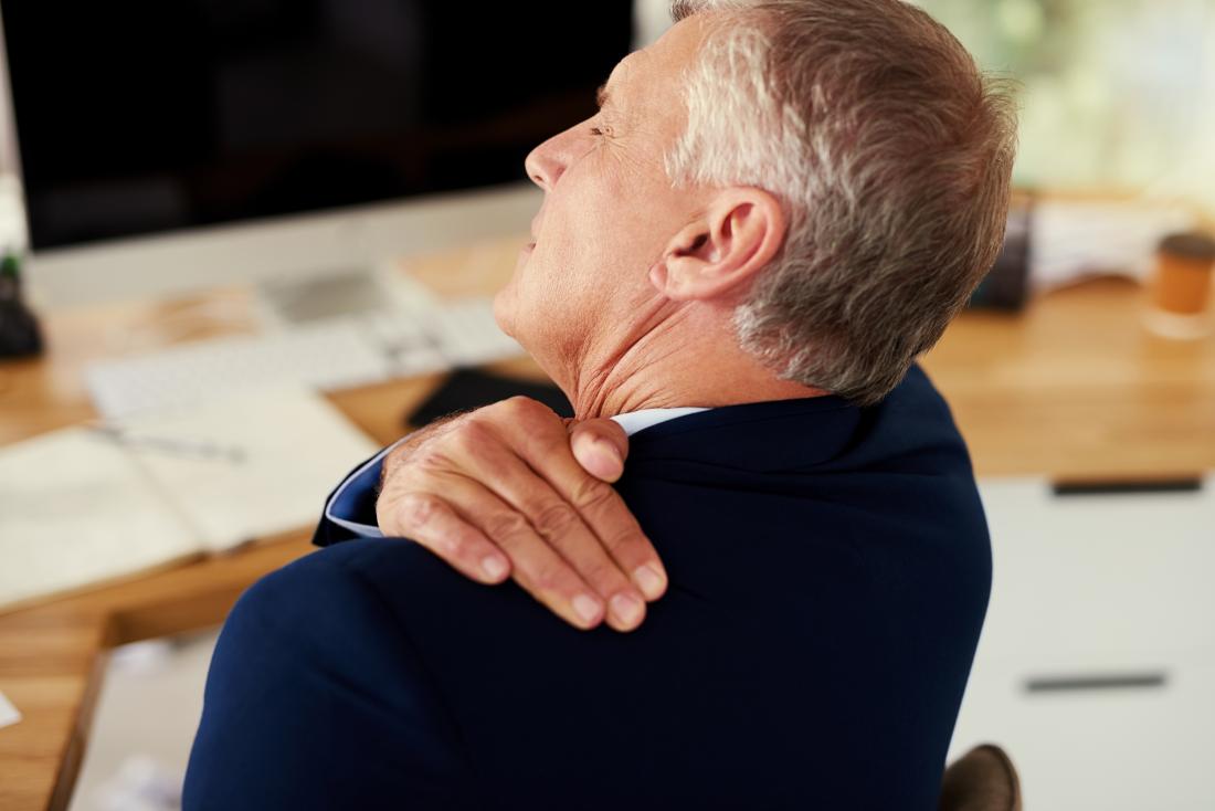 Businessman with shoulder bursitis holding top of shoulder and back in pain, sitting at desk at work.