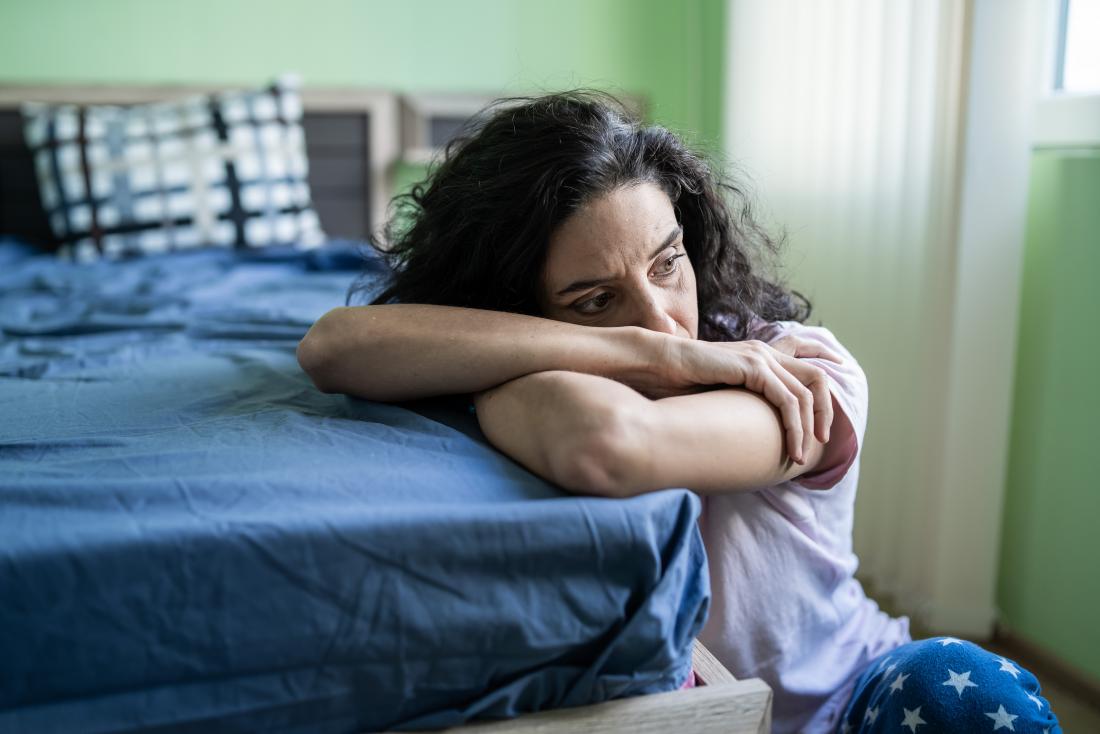 Woman sitting next to bed resting head on arms, suffering from insomnia and trouble sleeping, looking worried and stressed