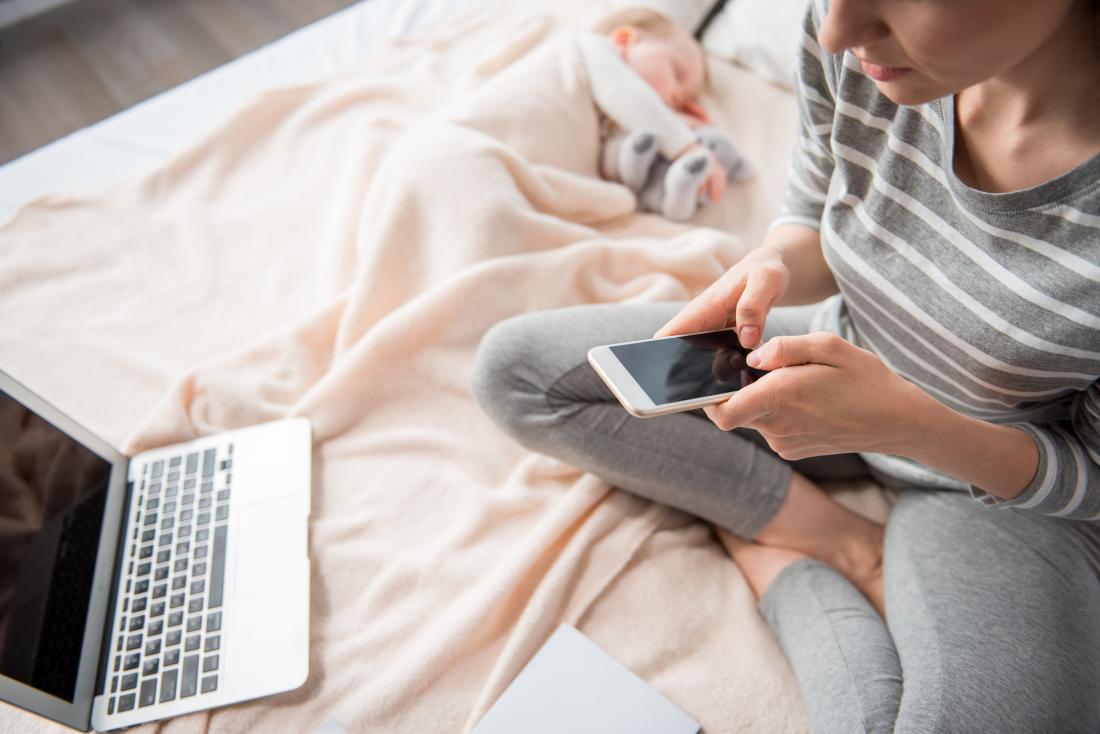 Mum on bed with sleeping baby and laptop while on phone