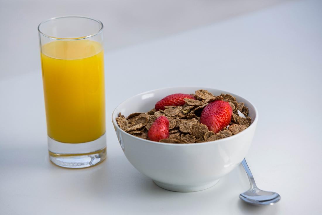 A bowl of bran flake cereal with strwberries on top, next to a glass of orange juice for breakfast.