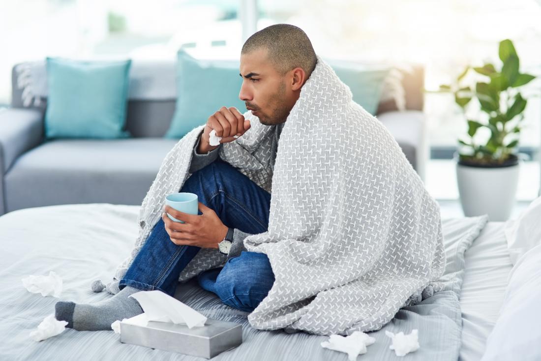 MAn with flu sitting under blanket coughing and drinking tea because of asthma complications.