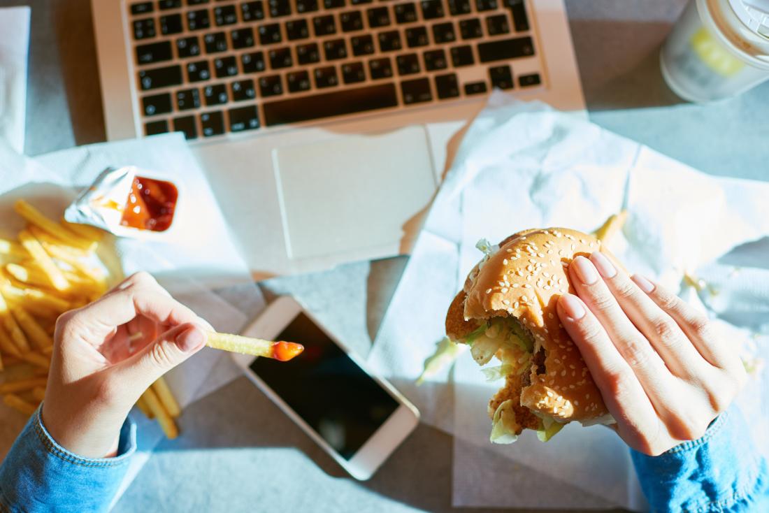 Person eating fast food burger and chips at their desk over their laptop and smartphone