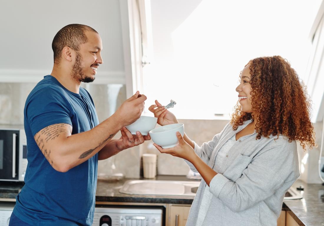 Couple eating breakfast cereal from bowls in kitchen.