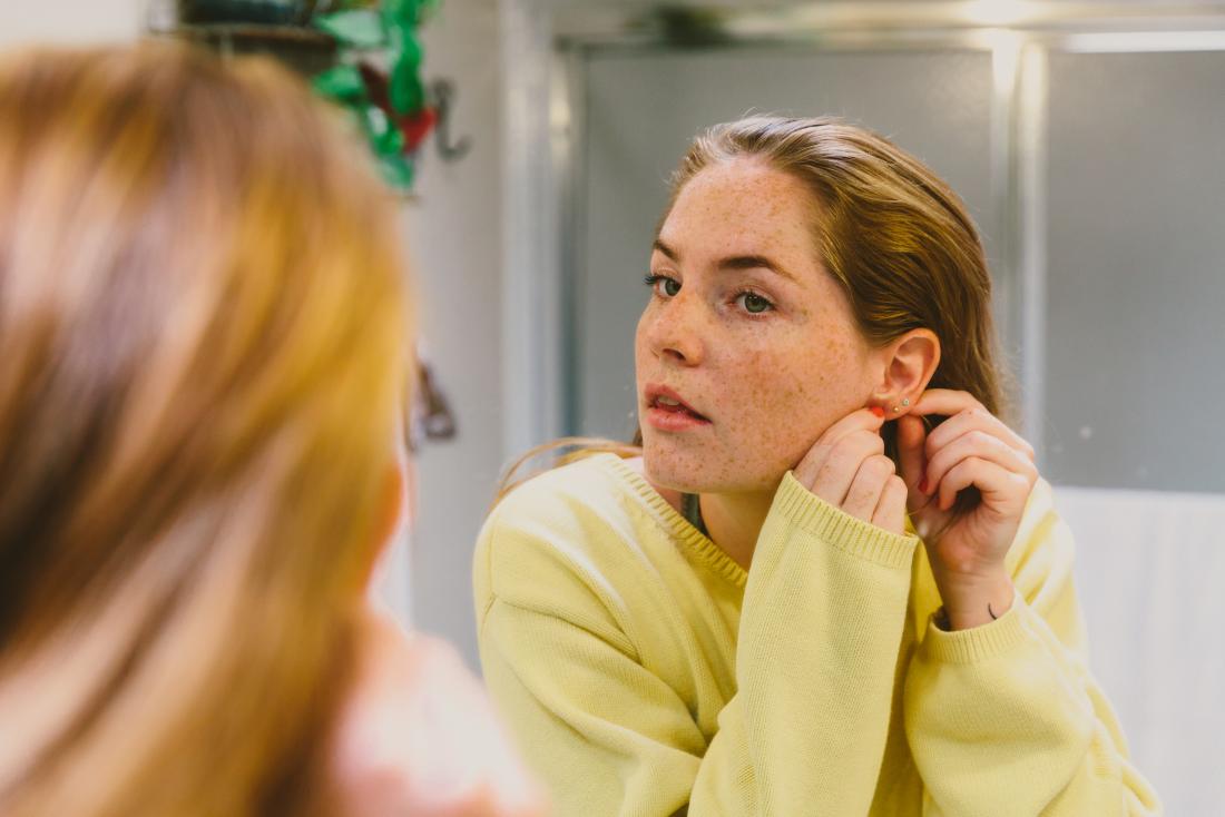 Woman with ear piercings looking in bathroom mirror touching behind ear.