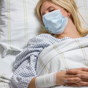 A woman with a bandaged arm rests in a hospital bed while wearing a surgical mask.