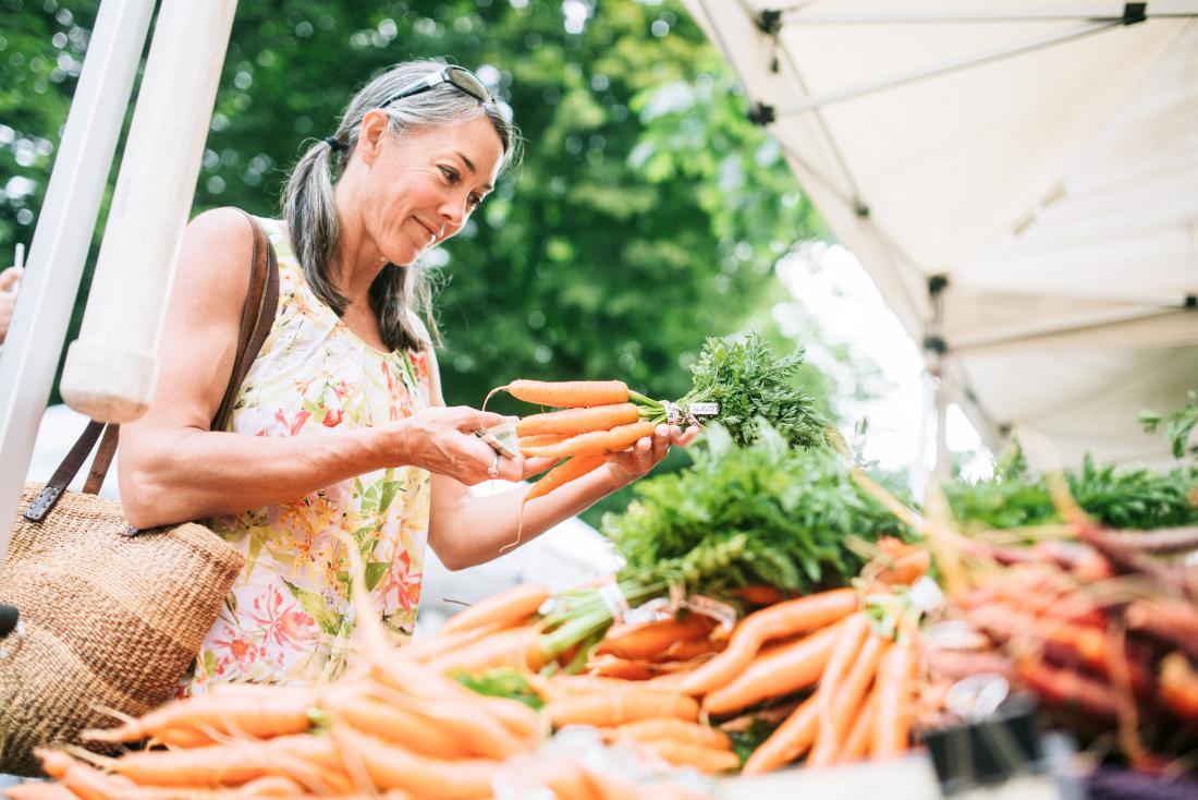 Woman holding carrots shopping for sustainable diet at outdoor farmers market.