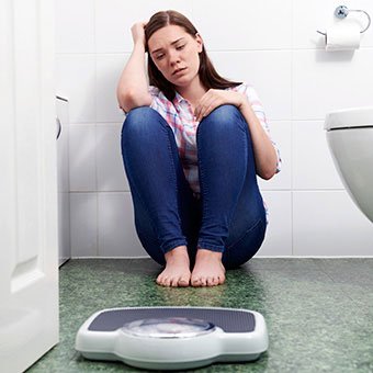 An upset female teenager sits on the floor of a bathroom next to a scale.